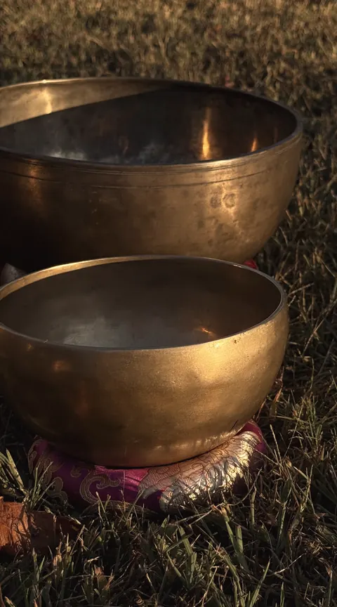 Two brass singing bowls on the grass at dusk, one resting on a red cushion